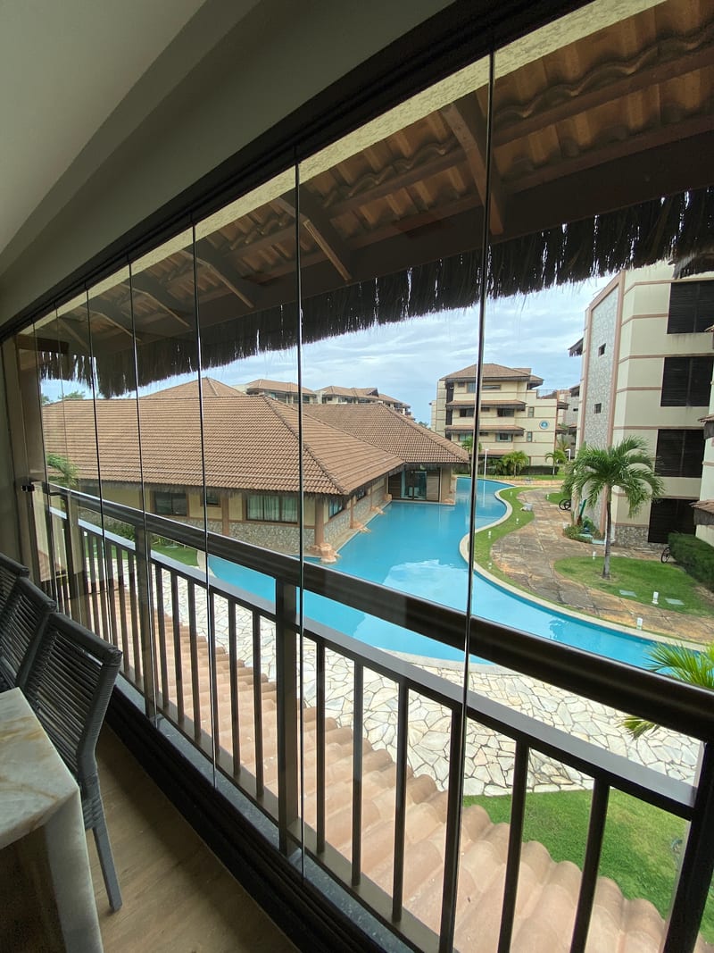 Varanda envidraçada em Fortaleza com vista para piscina e coqueiros, painéis de vidro recolhidos na lateral